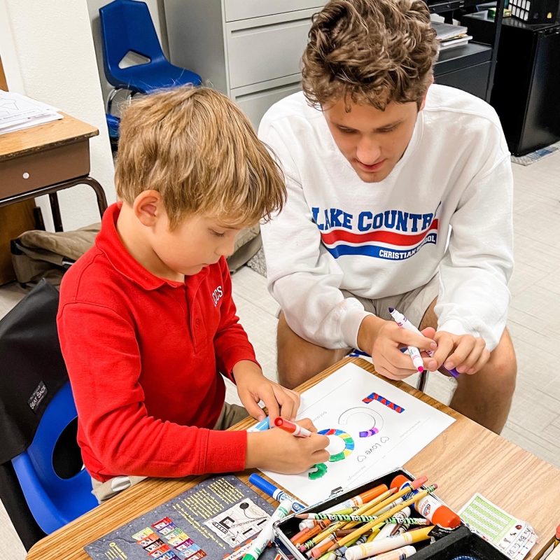 High school student working with an elementary student who is coloring a paper at a desk as part of the Bible Buddies program.