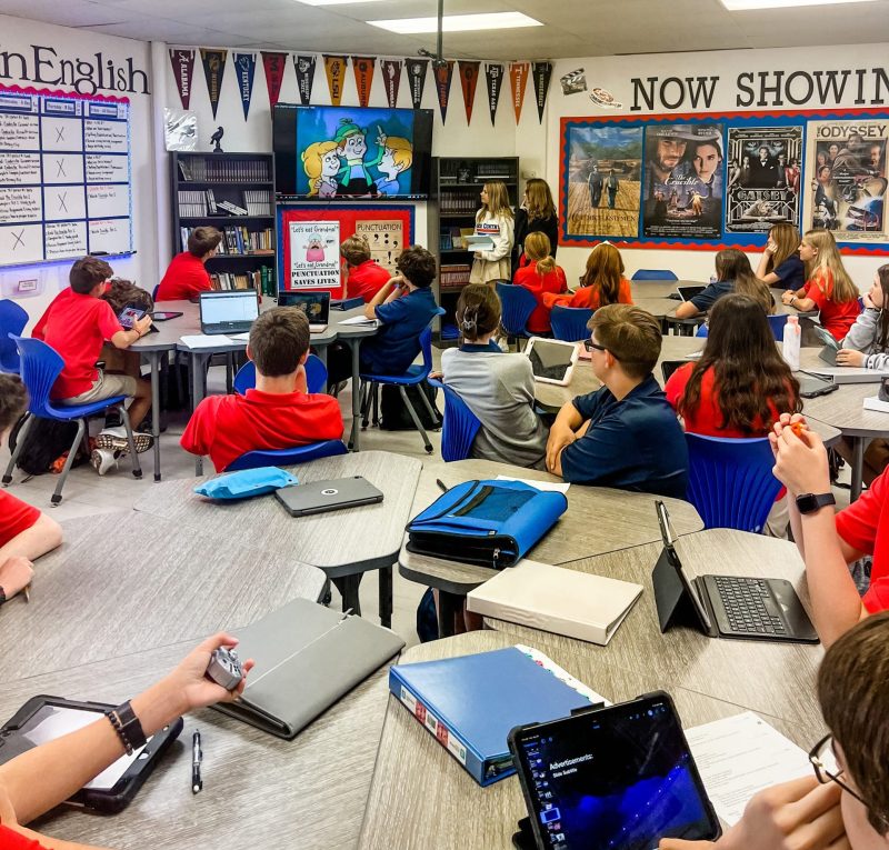 high school classroom with students at tables on iPads and two girls making a presentation at the tv on the far wall